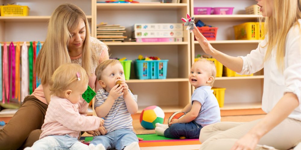 Group of babies in nursery. Nursery teacher and assistant play with kids on cosy rug
